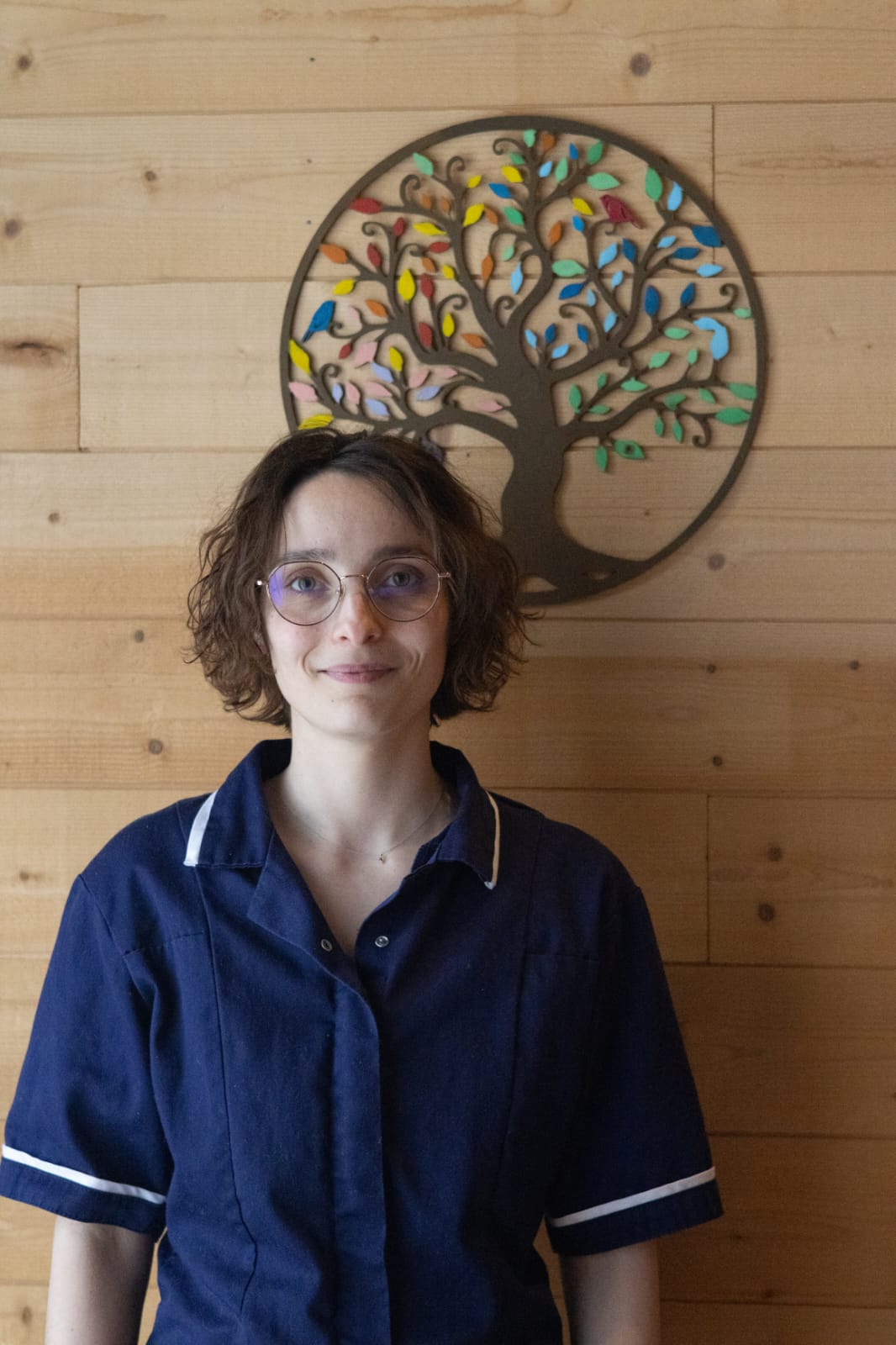 Léa Muller, réflexologue, souriante dans son cabinet Au Cœur des Sens devant l'arbre de vie.